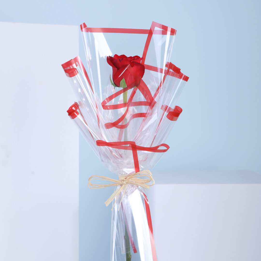 A single red rose wrapped in decorative clear and red cellophane tied with a raffia ribbon.